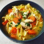 Close-up of a bowl of homemade chicken noodle soup featuring shredded chicken, thick carrot slices, and egg noodles.