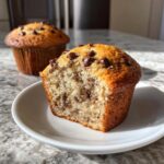 A chocolate chip banana muffins half, showing the moist interior texture, resting on a white plate.