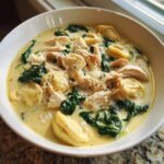 Close-up of a white bowl filled with creamy chicken tortellini soup, featuring shredded chicken and wilted spinach.