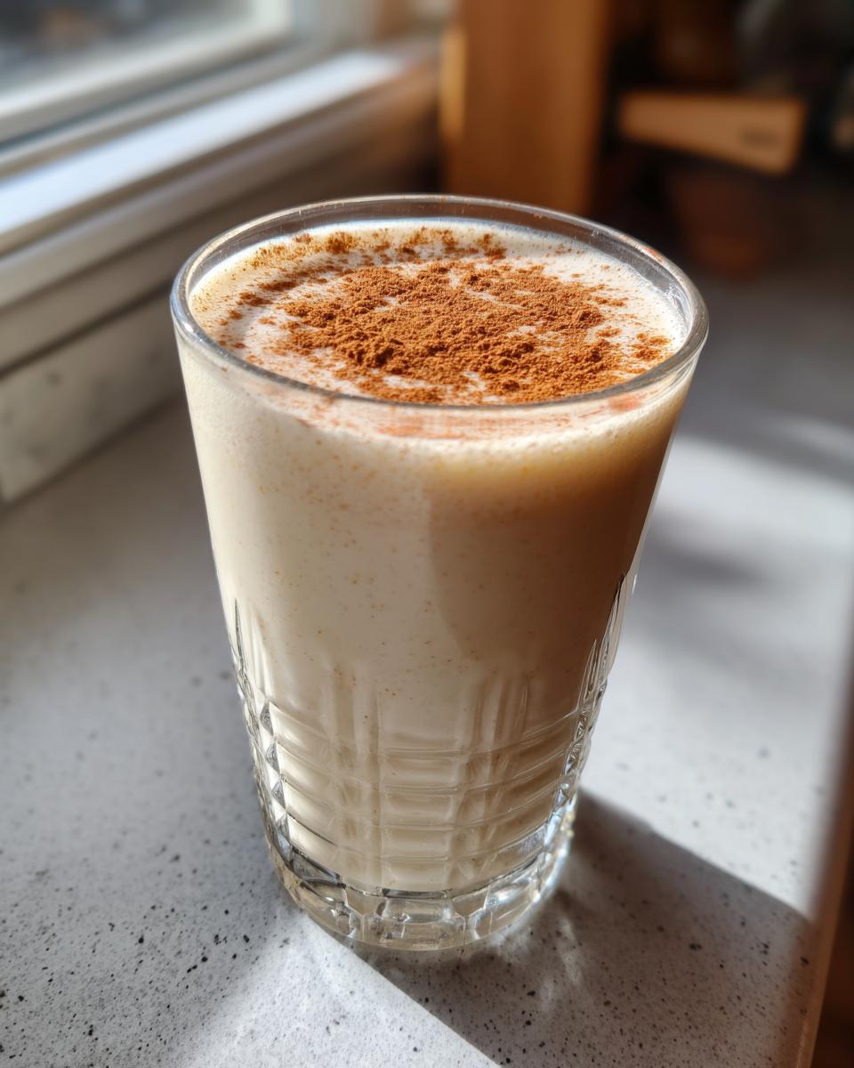 A glass of creamy, light-colored coquito topped generously with ground cinnamon powder.