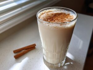 A tall glass filled with creamy coquito, topped generously with ground cinnamon, sitting next to two cinnamon sticks.