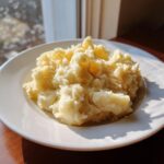 A generous serving of fluffy, creamy mashed potatoes recipe piled high on a white plate near a window.