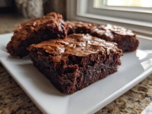 Close-up of fudgy brownies with shiny, cracked tops served on a white rectangular plate.