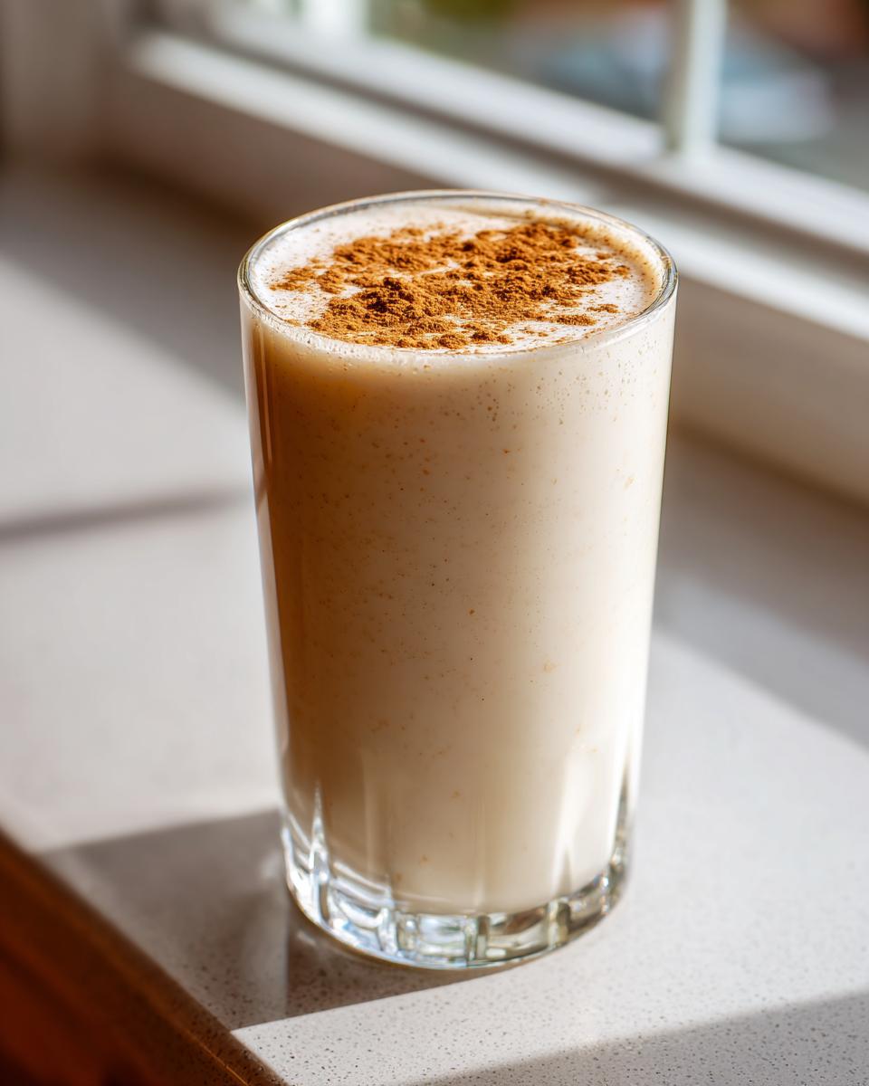 A tall glass filled with creamy, light-colored coquito topped generously with ground cinnamon.