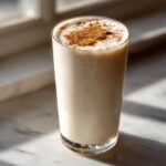 A tall glass of creamy, light-colored coquito garnished with ground cinnamon on top.