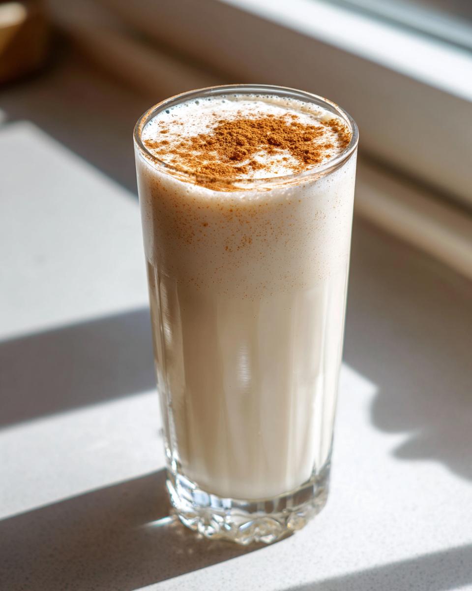 A tall glass filled with creamy, chilled coquito, dusted generously with ground cinnamon on top.