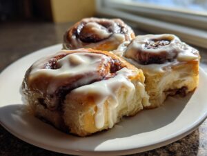 Three gooey cinnamon rolls topped with thick white frosting sitting on a white plate in bright sunlight.