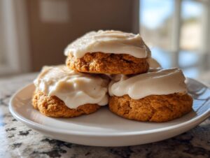A stack of three soft, orange pumpkin cookie treats topped generously with thick cream cheese frosting.