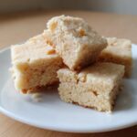 Close-up of four buttery, crumbly shortbread cookies squares stacked on a white plate.