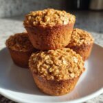 Four golden brown banana oatmeal muffins stacked on a white plate, highlighted by natural light.
