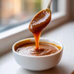 A spoonful of thick, glossy date caramel is drizzled back into a small white bowl.