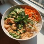 A close-up of a fresh spring roll salad bowl featuring shrimp, rice noodles, shredded carrots, cucumbers, and peanut sauce.