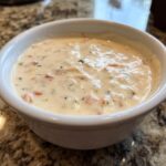 A close-up of a creamy, warm slow cooker cheese dip with visible chunks of tomato and herbs in a white bowl.