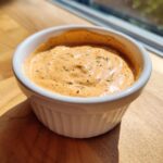 Close-up of creamy, orange-tinted smash burger sauce seasoned with herbs in a small white bowl.