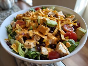 A close-up view of a hearty southwest salad bowl topped with chicken, avocado, tomatoes, and creamy orange dressing.