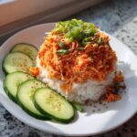 A serving of spicy crab sushi bowl featuring shredded spicy crab mixture over rice, garnished with green onions and sesame seeds, served with cucumber slices.