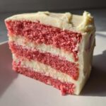 Close-up of a moist slice of pink layered strawberry cake with thick white cream cheese frosting.