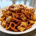 A close-up of homemade toffee chex mix recipe featuring glazed Chex cereal and pretzels piled high in a white bowl.
