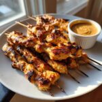A plate of grilled, golden brown chicken satay skewers served with a small bowl of peanut dipping sauce.