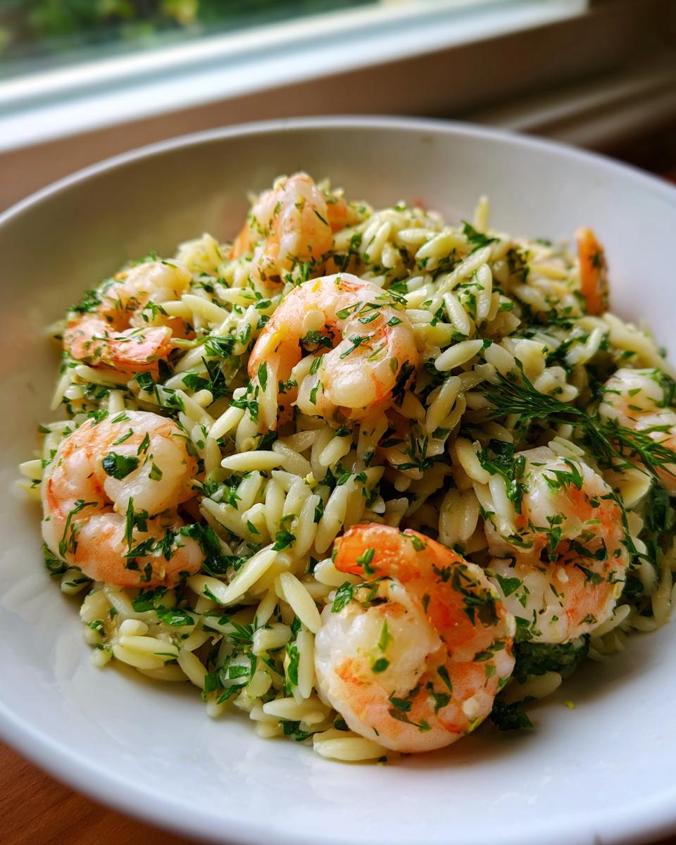 Close-up of a white bowl filled with amazing shrimp orzo salad, featuring plump pink shrimp and fresh green herbs.