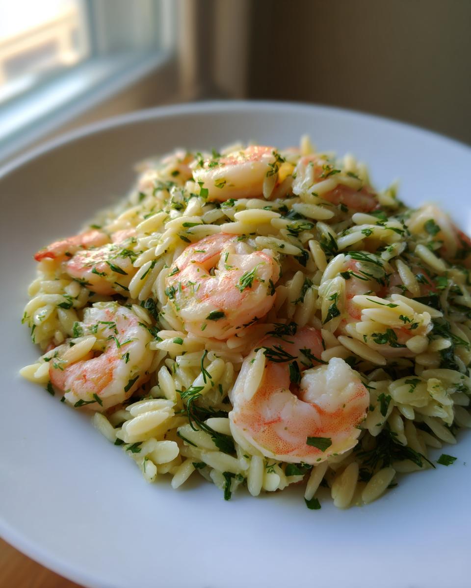 A close-up serving of amazing shrimp orzo salad mixed with fresh green herbs, presented on a white plate near a window.