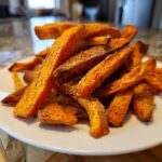 A close-up of a mound of perfectly seasoned sweet potato fries sprinkled with salt and pepper on a white plate.