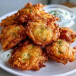 A close-up of several golden brown, crispy Amish onion fritters piled on a white plate with a side of white dipping sauce.