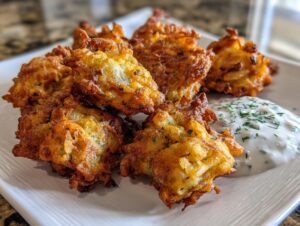 A serving of crispy, golden brown Amish onion fritters piled on a white plate next to a dollop of creamy dipping sauce topped with dill.