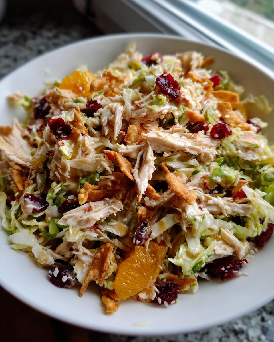 A close-up of a vibrant asian chicken cranberry salad in a white bowl, featuring shredded chicken, cabbage, dried cranberries, and mandarin orange slices.