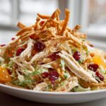 A close-up of a vibrant asian chicken cranberry salad featuring shredded chicken, dried cranberries, mandarin oranges, and crispy wonton strips.