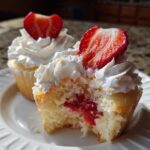 Close-up of a strawberry shortcake cupcake with a bite taken out, revealing strawberry filling and topped with whipped cream and a fresh strawberry slice.