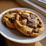 Close-up of two freshly baked brown butter chocolate chip cookies topped with melted chocolate chunks and sea salt.