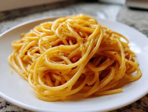 Close-up of a serving of glossy buttered spaghetti, lightly seasoned with red pepper flakes, on a white plate.