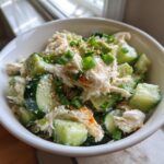 A close-up of a white bowl filled with california roll cucumber salad, featuring chunks of cucumber, imitation crab, and green onions.