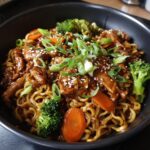 A close-up, appetizing shot of chicken ramen stir fry featuring glossy noodles, saucy chicken pieces, broccoli florets, and sliced carrots, topped with green onions and sesame seeds.