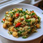 A close-up of a vibrant chickpea quinoa salad mixed with red peppers and fresh green herbs served in a white bowl.