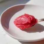 A spoonful of vibrant pink Cinnamon Rhubarb Butter resting on a white plate under bright light.