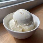 Two scoops of creamy white cottage cheese ice cream served in a small white bowl on a wooden surface.