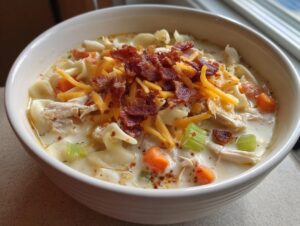 A close-up of a bowl of creamy crack chicken noodle soup topped with shredded cheese and crispy bacon bits.