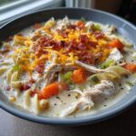 Close-up of a bowl of creamy crack chicken noodle soup with shredded chicken, egg noodles, carrots, and bacon topping.