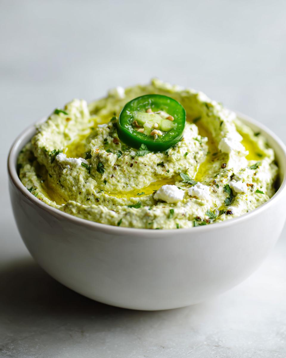 Close-up of a white bowl filled with vibrant green creamy jalapeño hummus, drizzled with olive oil and topped with a jalapeño slice.