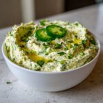 A white bowl filled with vibrant, creamy jalapeño hummus, drizzled with olive oil and topped with fresh cilantro and jalapeño slices.