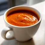 A close-up of rich, vibrant orange creamy tomato soup garnished with herbs in a white mug.