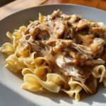 Close-up of shredded crockpot chicken and gravy generously poured over wide egg noodles on a white plate.