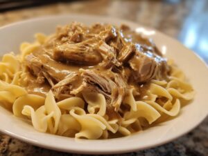A close-up of shredded crockpot chicken and gravy generously poured over egg noodles in a white bowl.