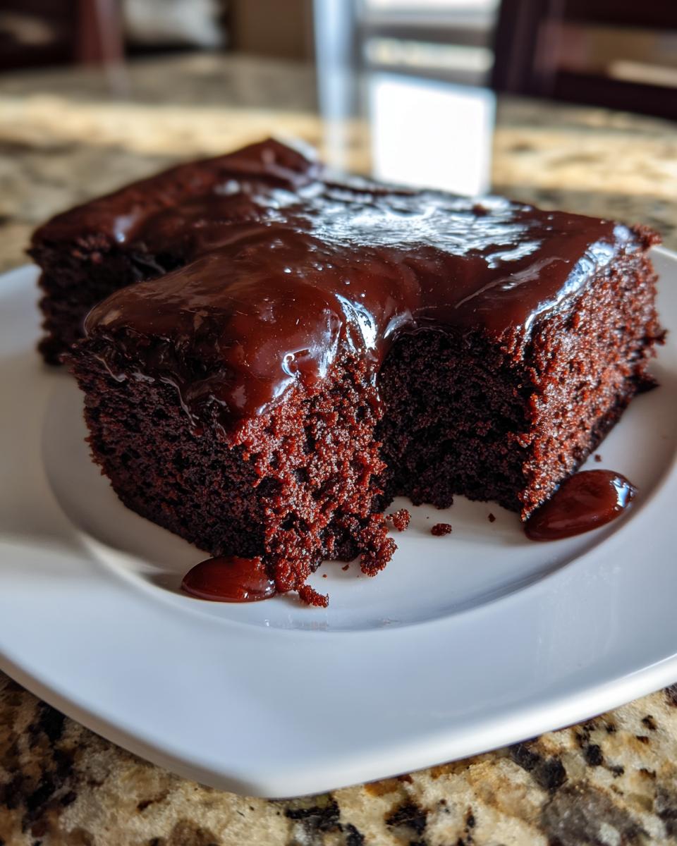 Close-up of a rich, dark slice of Dr Pepper Cake topped with glossy chocolate glaze on a white plate.