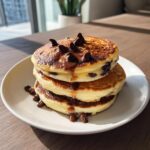 A tall stack of three fluffy chocolate chip pancakes drizzled with melted chocolate.