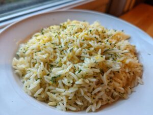 A close-up of fluffy, bright Greek rice seasoned with lemon zest and fresh green herbs served on a white plate.