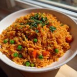 A close-up of fluffy, orange-hued mexican rice recipe served in a white bowl, mixed with green peas and carrots, topped with fresh cilantro.