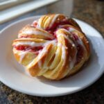 A single, beautifully shaped Geometric Rhubarb Cardamom Bun drizzled with white icing, resting on a white plate.